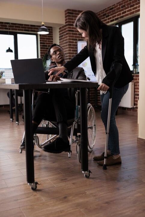 Fotografia colorida de um ambiente de trabalho com duas pessoas conversando: um homem em cadeira de rodas e uma mulher em pé, usando muleta.