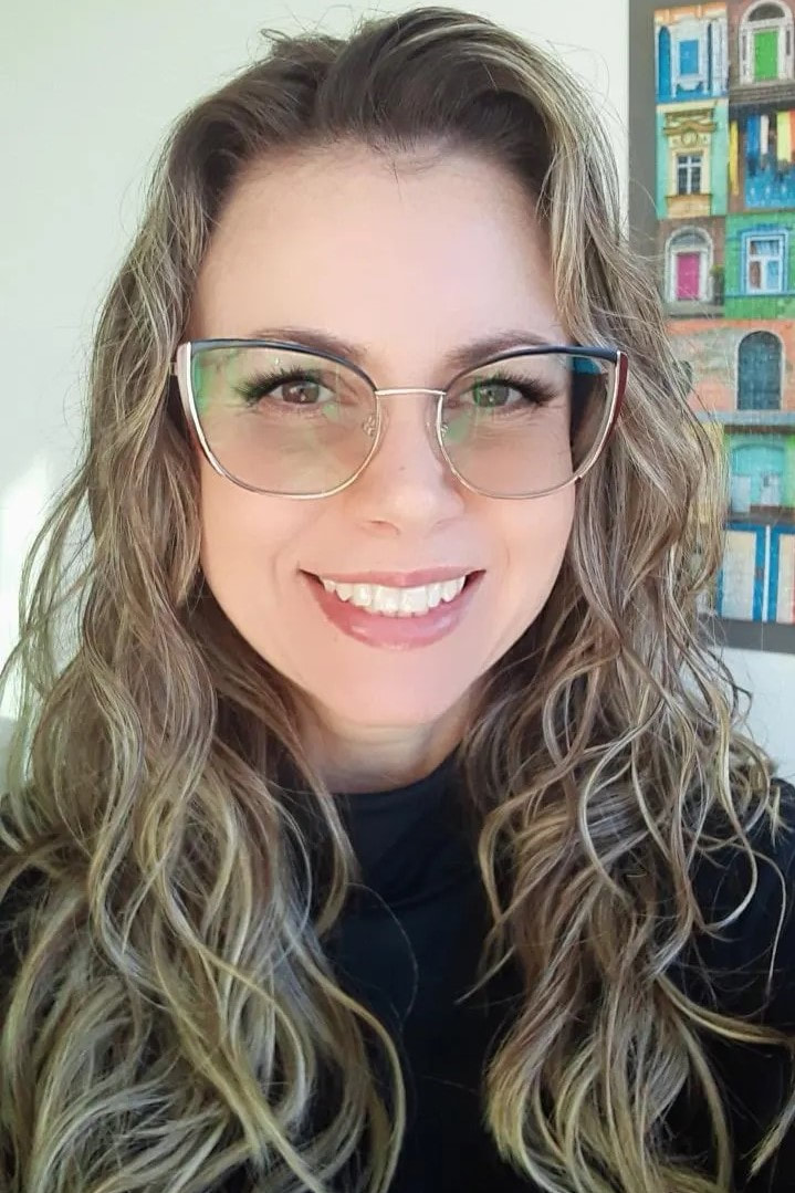 Fotografia colorida da Vanessa, uma mulher com pele branca, cabelo castanho claro longo e ondulado, olhos castanhos e óculos. Ela está sorrindo e veste uma blusa preta.