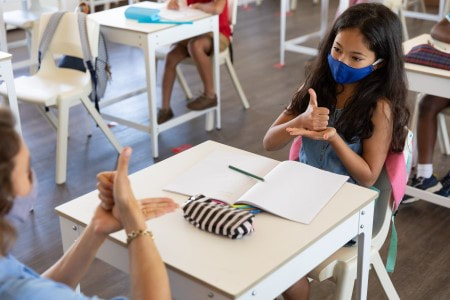 Fotografia colorida de uma criança e uma professora numa sala de aula. Elas estão conversando em língua de sinais.