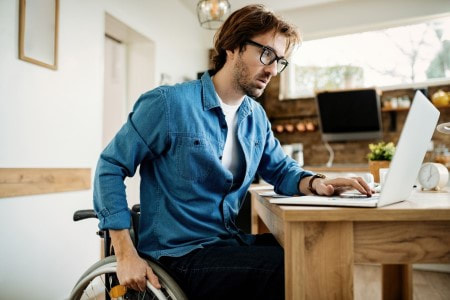 Fotografia colorida de um homem em cadeira de rodas trabalhando num computador.