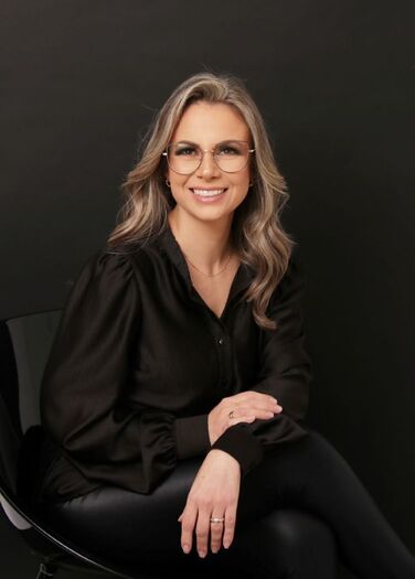 Fotografia colorida da Vanessa, uma mulher com pele branca, cabelo castanho claro longo e ondulado, olhos castanhos e óculos. Ela está vestindo camisa e calça pretas e está sentada numa cadeira preta. Está sorrindo, com o tronco levemente inclinado para frente, com as mãos repousando sobre as pernas cruzadas.