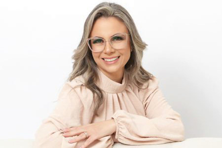 Fotografia colorida da Vanessa, uma mulher com pele branca, cabelo castanho claro longo e ondulado, olhos castanhos e óculos. Ela está sorrindo e veste uma blusa bege.