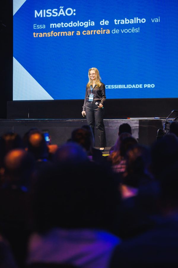 Fotografia colorida da Vanessa no palco durante uma palestra. Ela veste camisa e calça pretas e segura um microfone na mão esquerda.