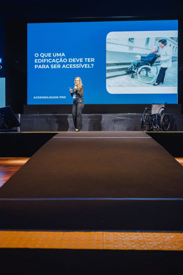 Fotografia colorida da Vanessa no palco durante uma palestra. Ela veste camisa e calça pretas e segura um microfone na mão esquerda.