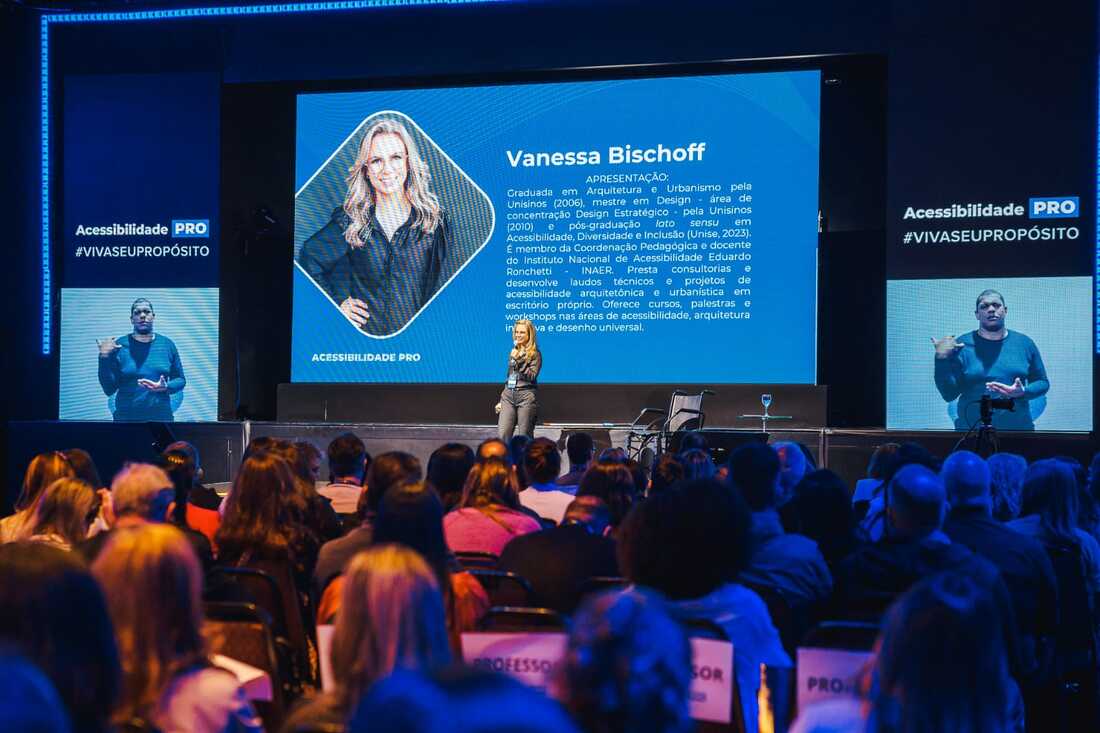 Fotografia colorida da Vanessa no palco durante uma palestra. Ela veste camisa e calça pretas e segura um microfone na mão esquerda.
